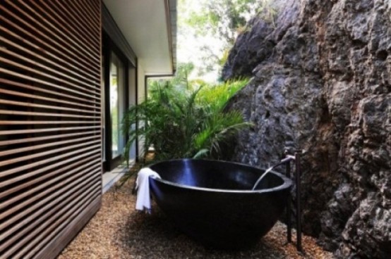 a tropical outdoor bathroom with a stone wall, a metal bathtub and potted greenery
