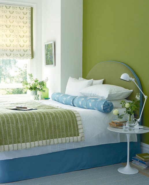 a light green bedroom with a matching bed with white, green and blue bedding, a printed curtain and art and a cool table lamp