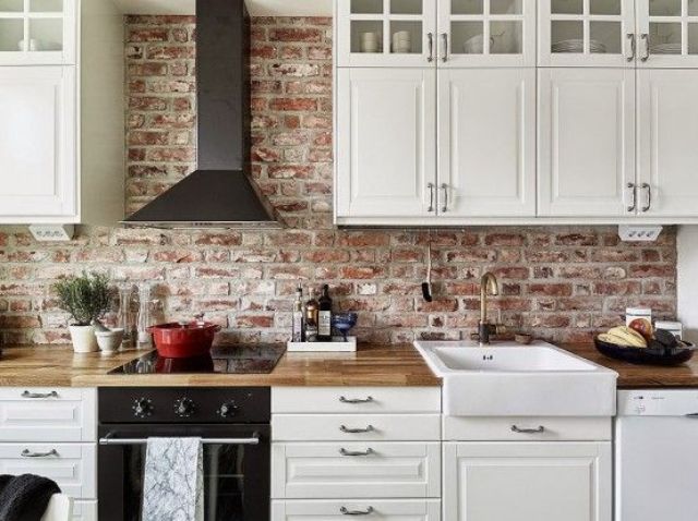 A small and pretty Scandinavian kitchen with shaker cabinets, butcherblock countertops, a black hood, a built in cooker and a red brick backsplash that adds interest and eye catchiness to the space