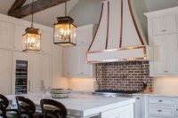 a vintage white kitchen with shaker style cabinets, built-in lights and a large hood with copper detailing, a red brick backsplash for a touch of color and more interest, pendant lamps and dark-stained stools