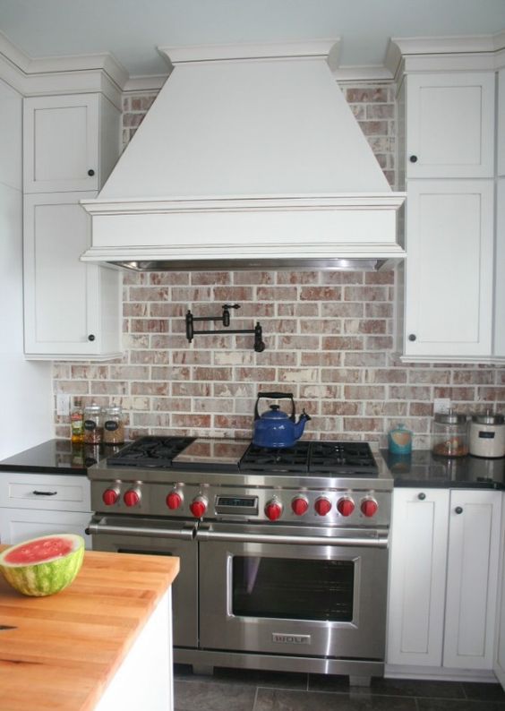 a white kitchen with shaker style cabinets, a large hood and a whitewashed red brick backsplash that adds interest, texture and color to the room