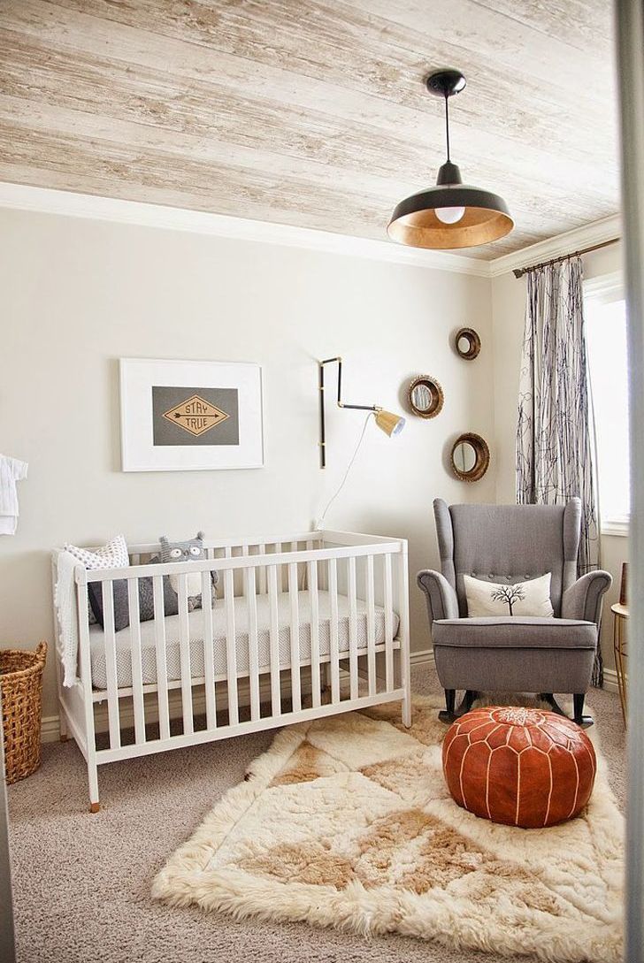 A welcoming mid century modern nursery in neutrals, with a white crib, a grey rocker chair, layered rugs, a leather pouf and some artwork