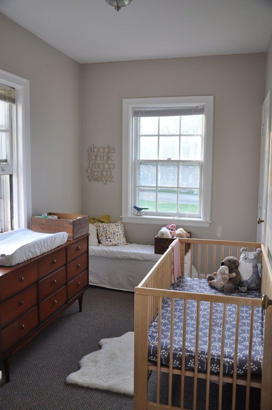 A tiny neutral nursery with a daybed, a mid century modern dresser that is a changing table, a crib with neutral bedding and layered rugs is amazing
