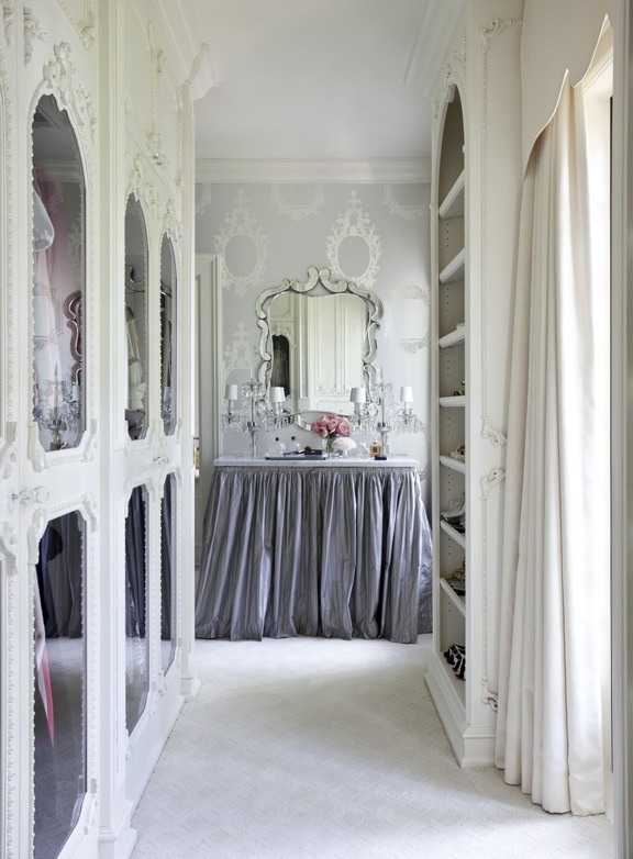 A delicate and girlish walk in closet with white ornated frame and mirror doors plus an open storage unit and a vanity with a lilac skirt and a vintage mirror