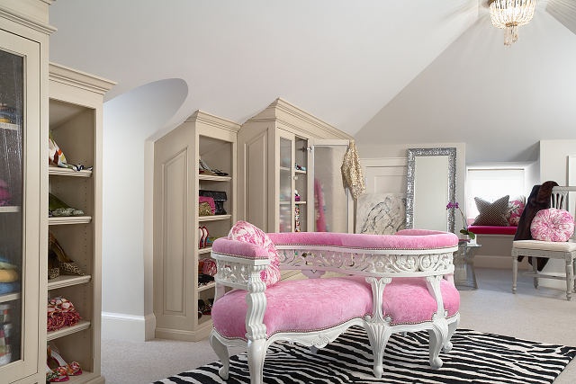 A pretty feminine walk in closet with built in storage units, a pink curved upholstered bench, a printed rug and some pink touches around