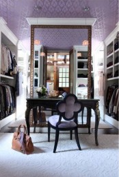 a vintage feminine walk-in closet in lilc, with a lilac print wallpaper on the ceiling, open storage units around, a large mirror and a black vanity plus a lilac chair