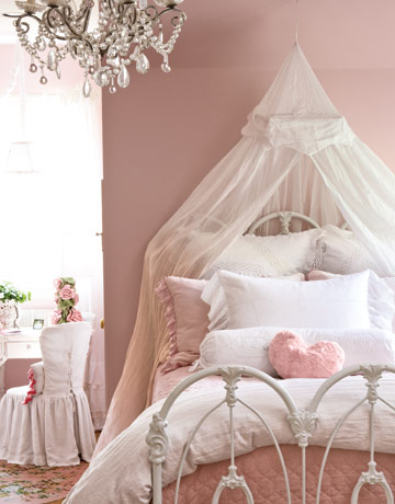 A dusty pink princess style room with an accent wall, a metal bed with pink and white bedding and a semi sheer canopy, a crystal chandelier, a desk and a metal chair
