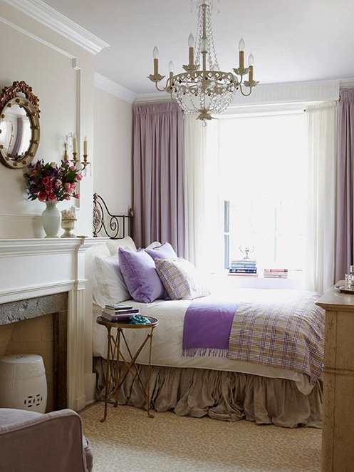 A French country bedroom with a non working fireplace, a forged bed, some plaid and lavender bedding, a crystal chandelier and a mirror