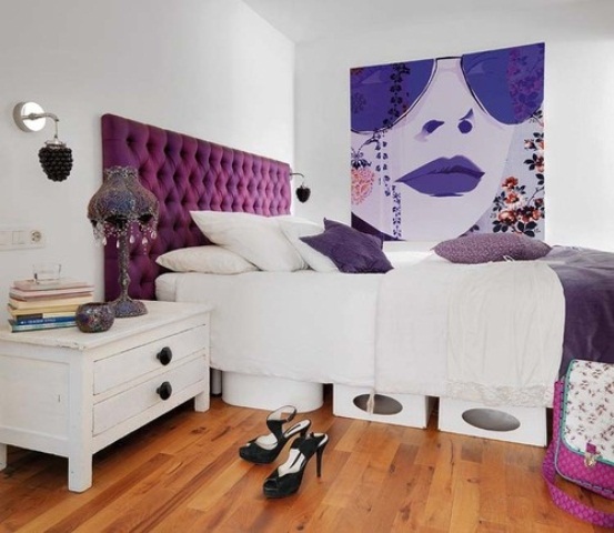 a white bedroom with a purple bed, purple pillows and an artwork, a vintage table lamp with crystals and some storage boxes