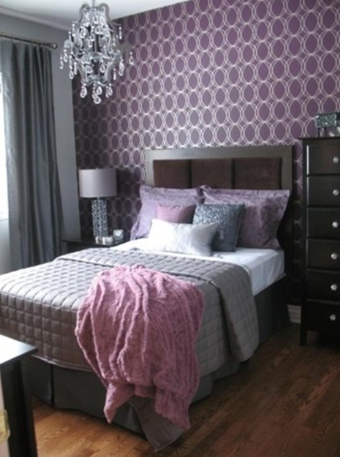 a stylish and refined bedroom with purple printed wallpaper, dark furniture, a crystal chandelier, grey and purple bedding and a wooden floor