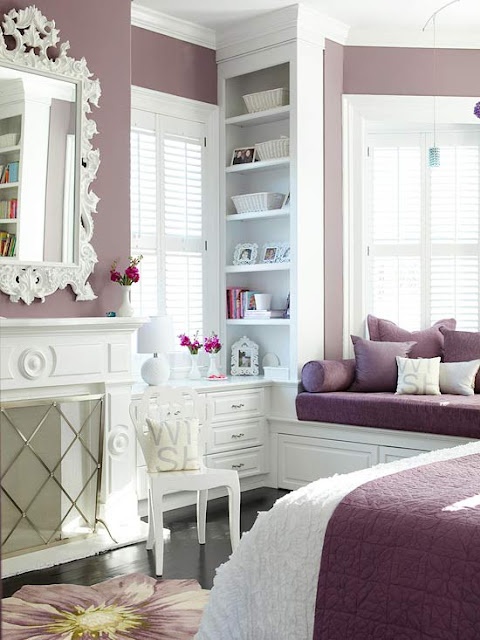 A purple and white bedroom with purple walls and vintage white furniture plus a non working fireplace