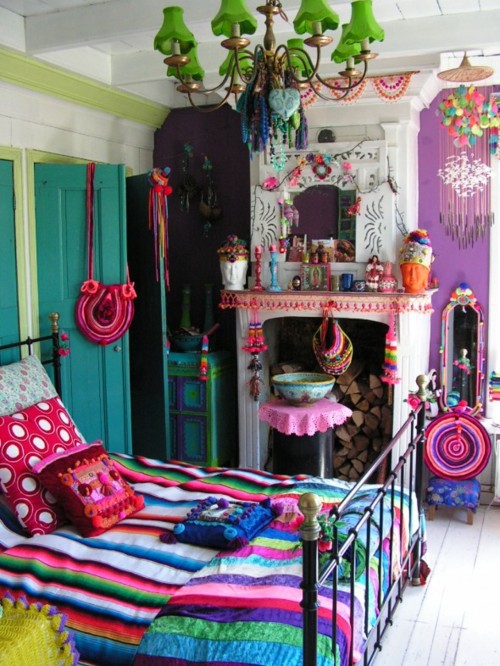 a colorful boho bedroom with a fireplace, a purple wall, a teal wardrobe, a forged bed with colorful and printed bedding, a green chandelier and lots of colorful accessories and art