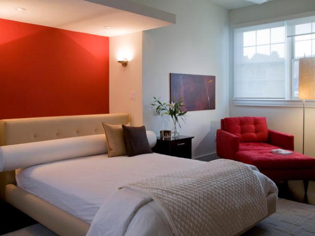 A modern neutral bedroom with a red accent wall, a bed with neutral bedding, a red chair with a footrest, lamps and sconces