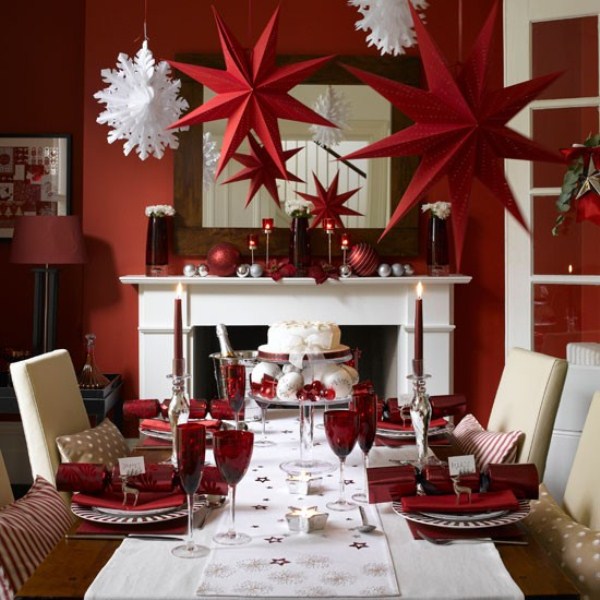 A stylish red and white Christmas dining room with a non working fireplace, a mirror over it, a table with neutral chairs, red and white paper stars hanging over the tabl and some red tableware