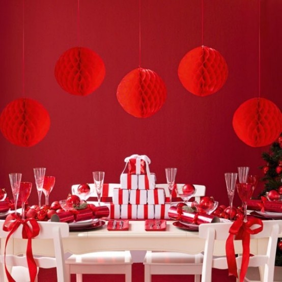 a red Christmas party space with a red accent wall, red paper pompoms, a table with red ornaments, red glasses and a stack of gift boxes with red ribbon