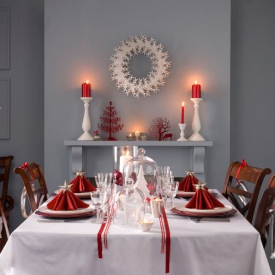 a red and white Christmas tablescape with white linens and red napkins shaped as Christmas trees and topped with stars, with a white cloche centerpiece and candles is a chic idea