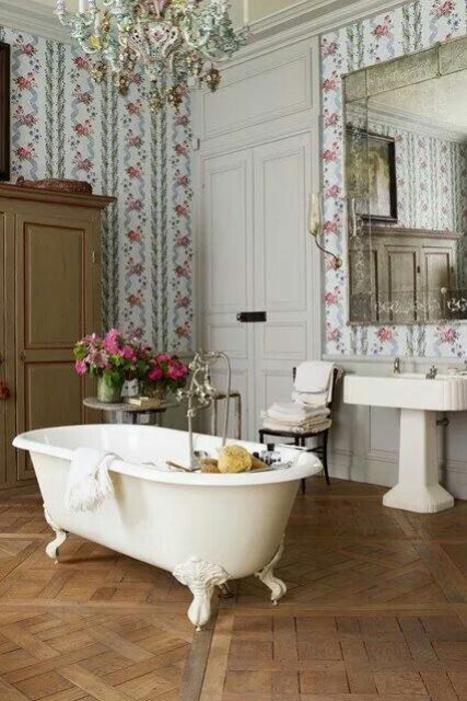 A lovely vintage bathroom with floral wallpaper, a wardrobe a white clawfoot bathtub, a free standing sink, an oversized mirror and a gorgeous parquet floor
