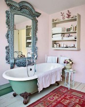 a vintage pink bathroom with a green clawfoot bathtub, with a shelving unit on the wall, a large mirror in an ornated frame, a bright rug and shabby chic furniture