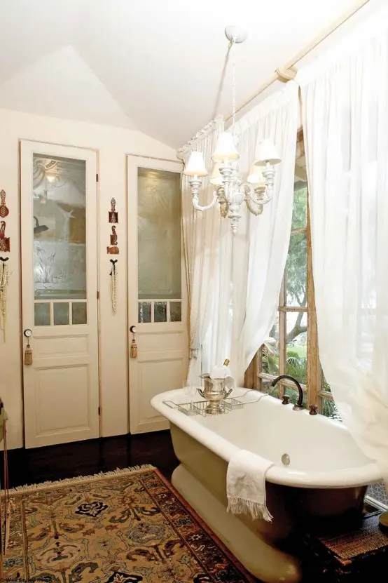 a chic vintage bathroom with neutral storage units built-in, a tan free-standing bathtub, a boho rug, a vintage white chandelier and white curtains