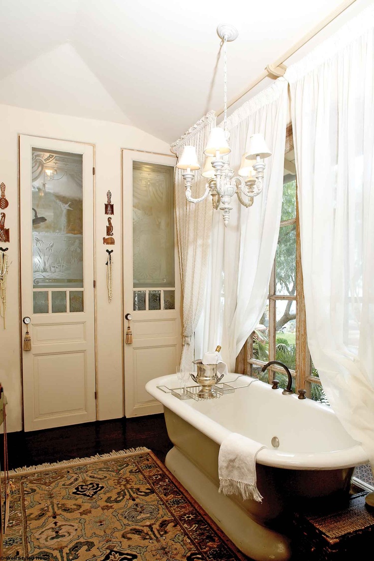 A chic vintage bathroom with neutral storage units built in, a tan free standing bathtub, a boho rug, a vintage white chandelier and white curtains