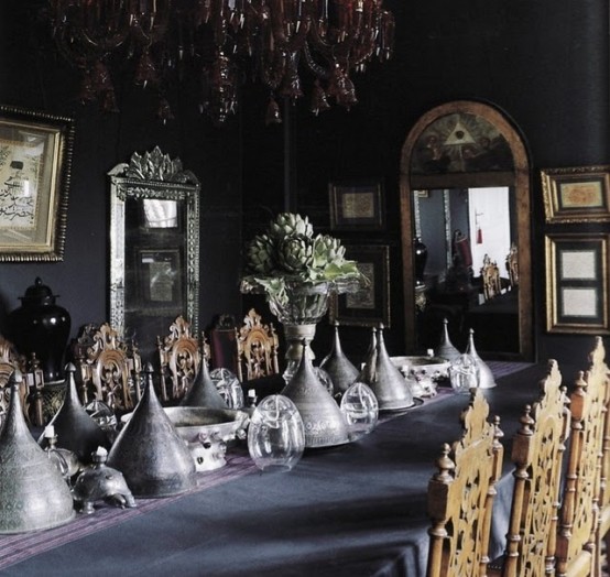 a beautiful and moody Gothic dining room with black walls, a gorgeous red chandelier, mirrors and artworks, a long table with cones and carved wooden chairs