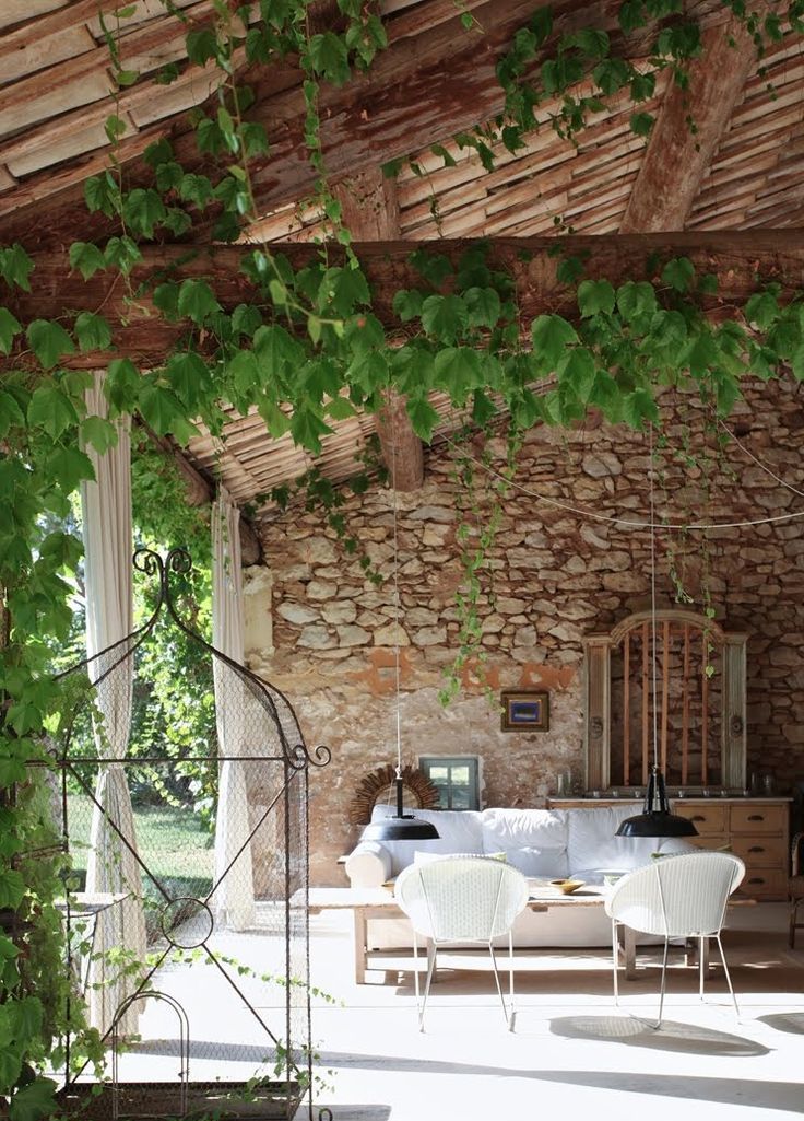 A Provence outdoor indoor space with lots of vines, a white sofa, a coffee table and some chairs, a sideboard and vintage cages for decor