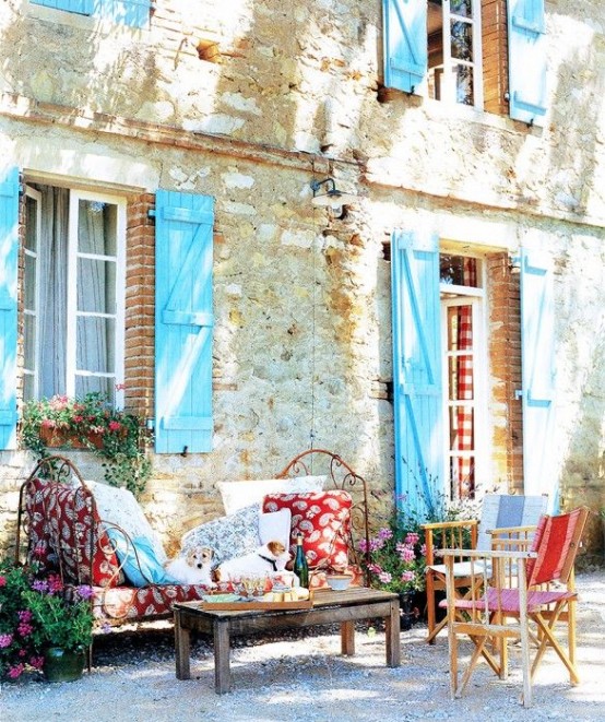 a bright and colorful Provence terrace with a forged bed with colorful cushions and pillows, a stained table and some neutral chairs with colorful pillows and cushions