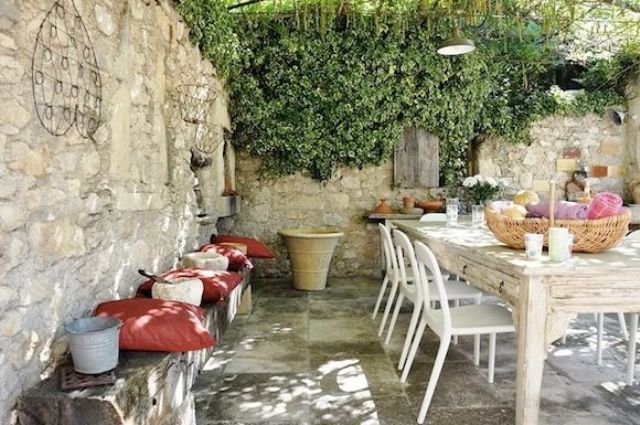 A neutral Provence terrace with greenery forming a roof, a whitewashed table and white chairs, a wooden bench with pillows and some planters and baskets