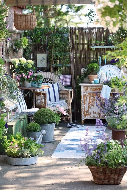 A small Provence inspired terrace with a wicker chair and a shabby chic cabinet, lots of potted greenery and blooms and some watering cans