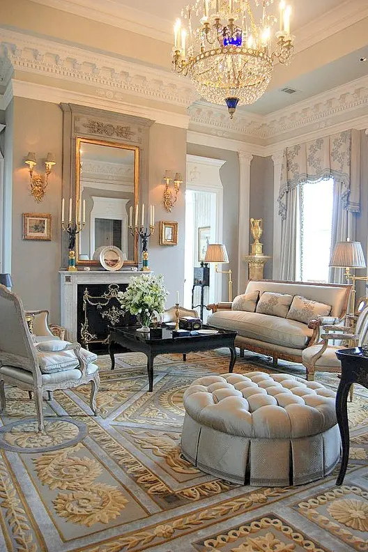 a sophisticated light grey and gold room with gorgeous molding on the ceiling, refined furniture, a large fireplace, a large mirror and a chic crystal chandelier