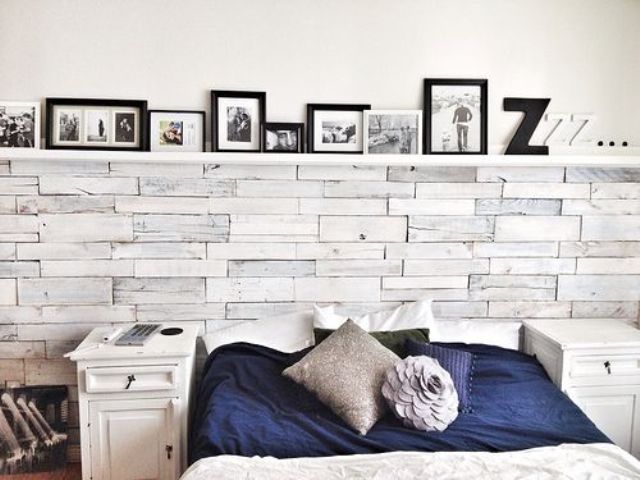 A modern rustic bedroom with a whitewashed wooden wall, white nightstands, a ledge with lots of artworks and contrasting bedding