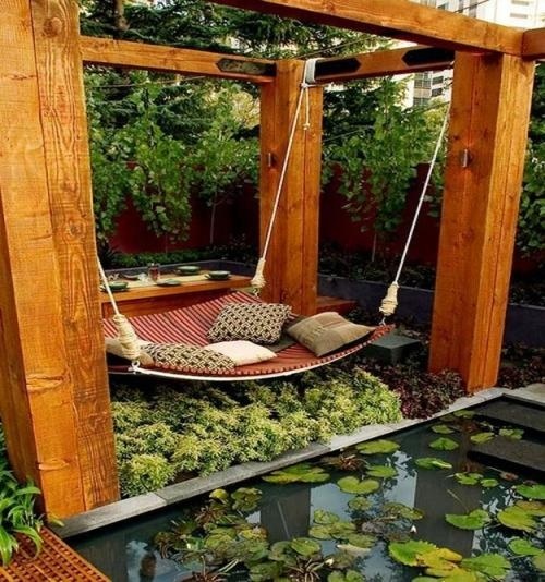 a zen relaxing space with a wooden gazebo and a hanging bed of pallets with some pillows next to a pond is very peaceful