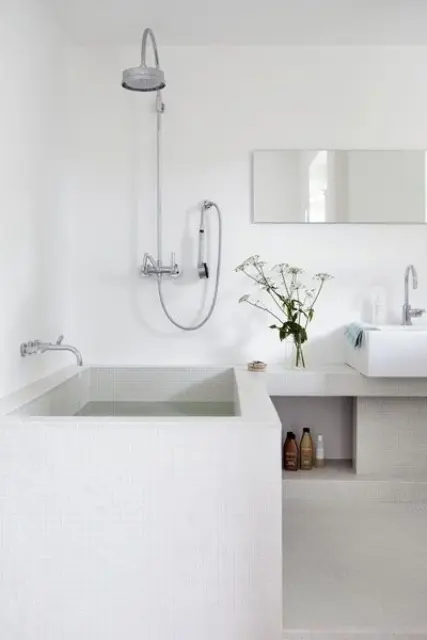 A minimalist Scandinavian bathroom in white and off white with a tile clad bathtub and niches for storage
