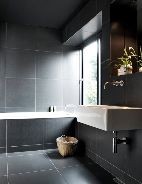 A moody Nordic bathroom fully clad with large grey tiles, with a wall mounted sink and a tub clad with tiles
