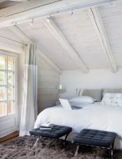 a white attic bedroom with a bed, black leather stools and a brown rug for a drama and a contrast