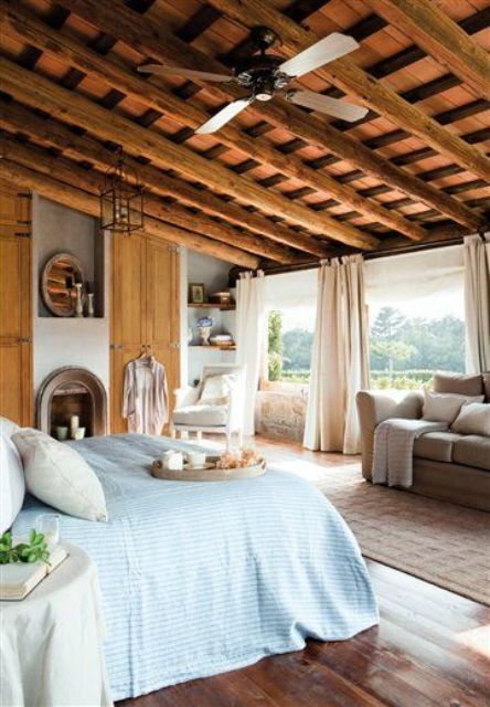 A rustic Provence bedroom with large windows, a faux fireplace with candles, built in shelves, neutral furniture, a bed with blue bedding