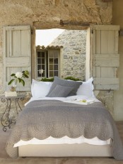 a neutral Provence bedroom with stone walls, a neutral bed with grey and white bedding, wooden shutters and a delicate nightstand and blooms
