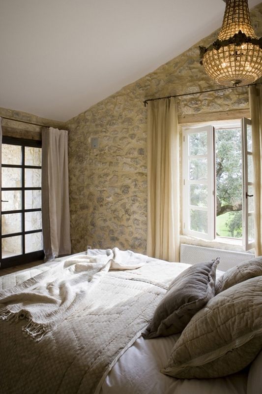 a Provence bedroom with a stone accent wall, a bed with neutral bedding, a crystal chandelier and much natural light