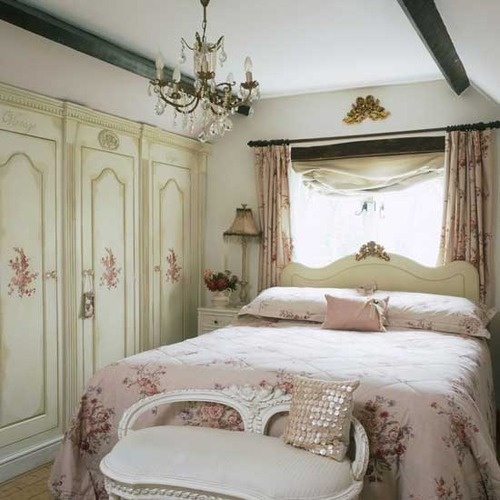 a refined vintage bedroom neutral storage units with floral patterns, a beautiful bed and a bench, a crystal chandelier and pink floral linens is lovely and chic