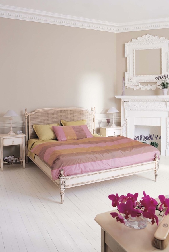 A vintage inspired feminine bedroom with grey walls, a carved wooden bed, a faux fireplace, picture frames and blooms