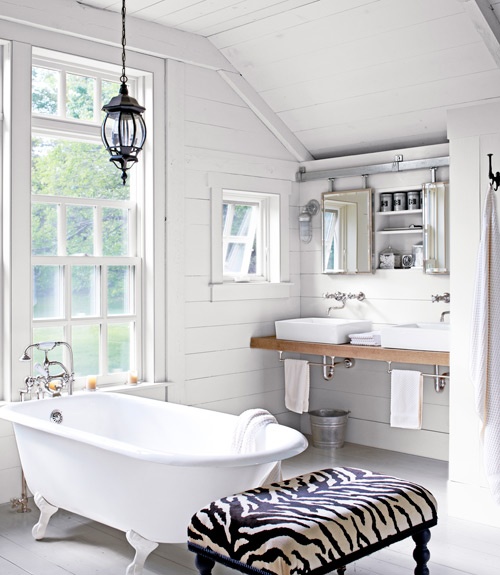 a white barn bathroom with wooden plank clad walls, a vintage lamp, a clawfoot tub, a printed ottoman and two mirrors