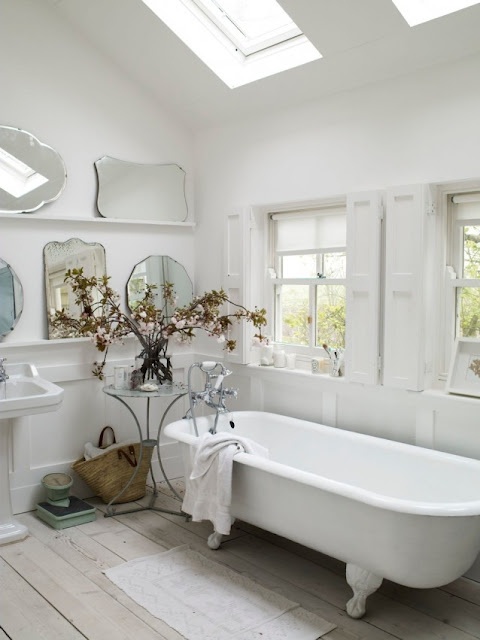 an elegant white bathroom with skylights and windows, shutters, a clawfoot tub, an arrangement of mirrors and some dried blooms