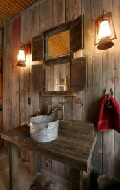 a barn bathroom clad with weathered wood, with mini windows with shutters, a metal tub sink and vintage lanterns on the wall