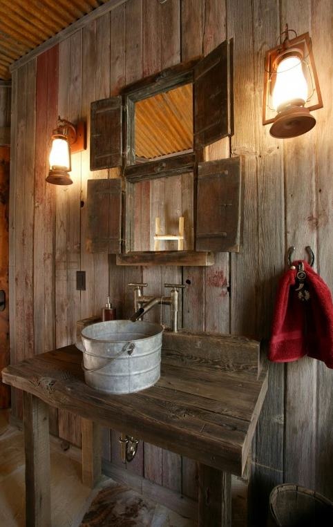 a barn bathroom clad with weathered wood, with mini windows with shutters, a metal tub sink and vintage lanterns on the wall