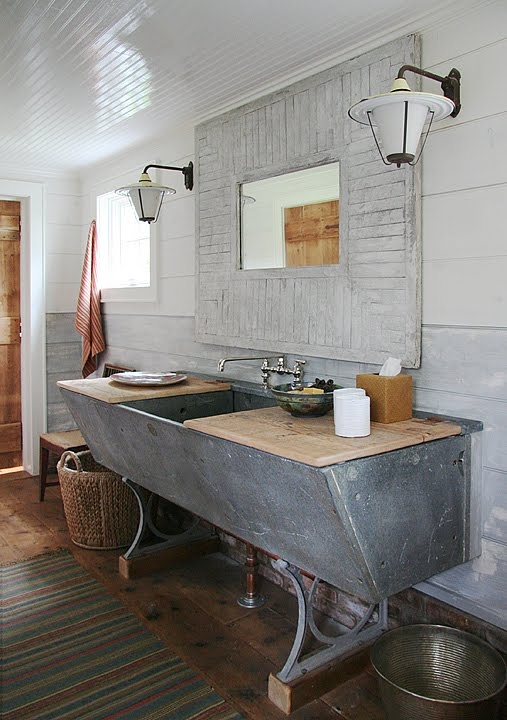 a shabby rustic bathroom with a large concrete sink, a whitewashed shabby chic mirror, baskets and rugs