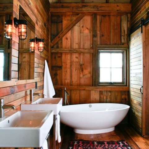 a rustic cabin bathroom fully clad with reclaimed and weathered wood, with white wall-mounted sinks, a free-standing tub and mirrors plus wall sconces
