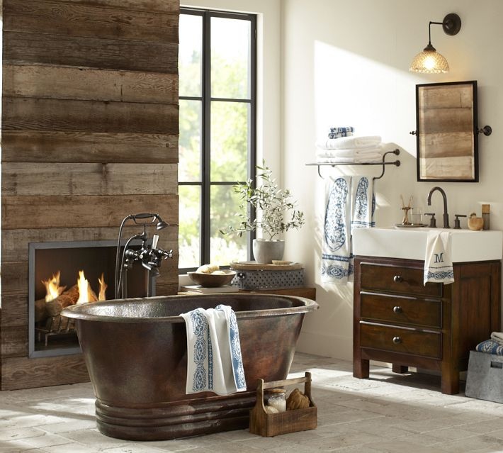 A rustic barn bathroom with a reclaimed wooden wall, wooden furniture, a built in fireplace and a vintage metal bathtub