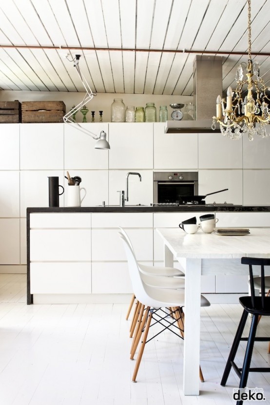 a Nordic off-white kitchen with a wooden ceiling, white sleek cabinets and a black countertop, a vintage chandelier and white chairs on wooden legs