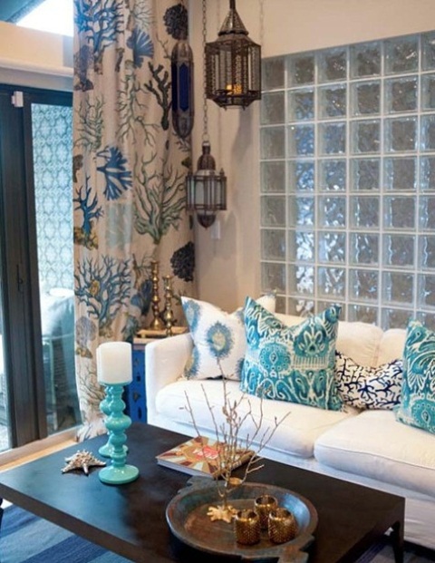 A sea inspired living room with a light blue glass wall, a white sofa with printed pillows, a printed curtain and Moroccan lanterns