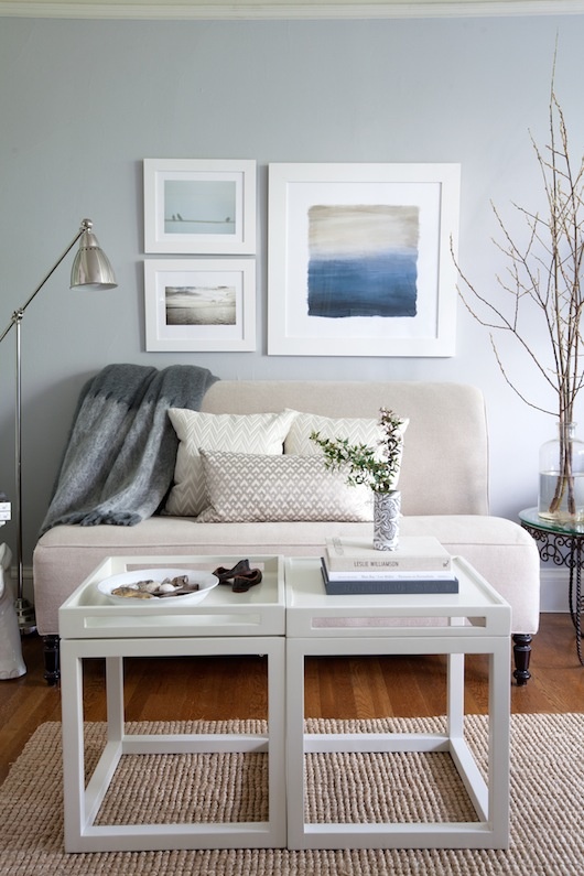 A neutral and light grey beach living room with a small vintage seat, a couple of tables and a sea inspired gallery wall